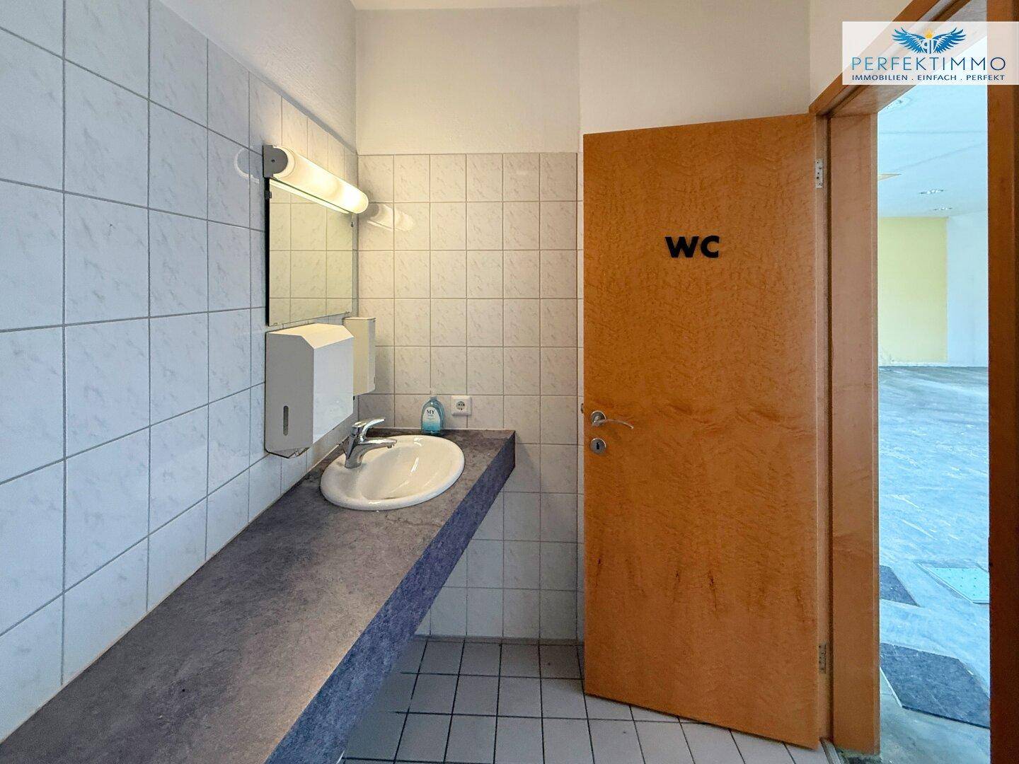 Waschbereich in einer Toilette mit Waschbecken, Spiegel und Seifenspender an gefliesten Wänden.