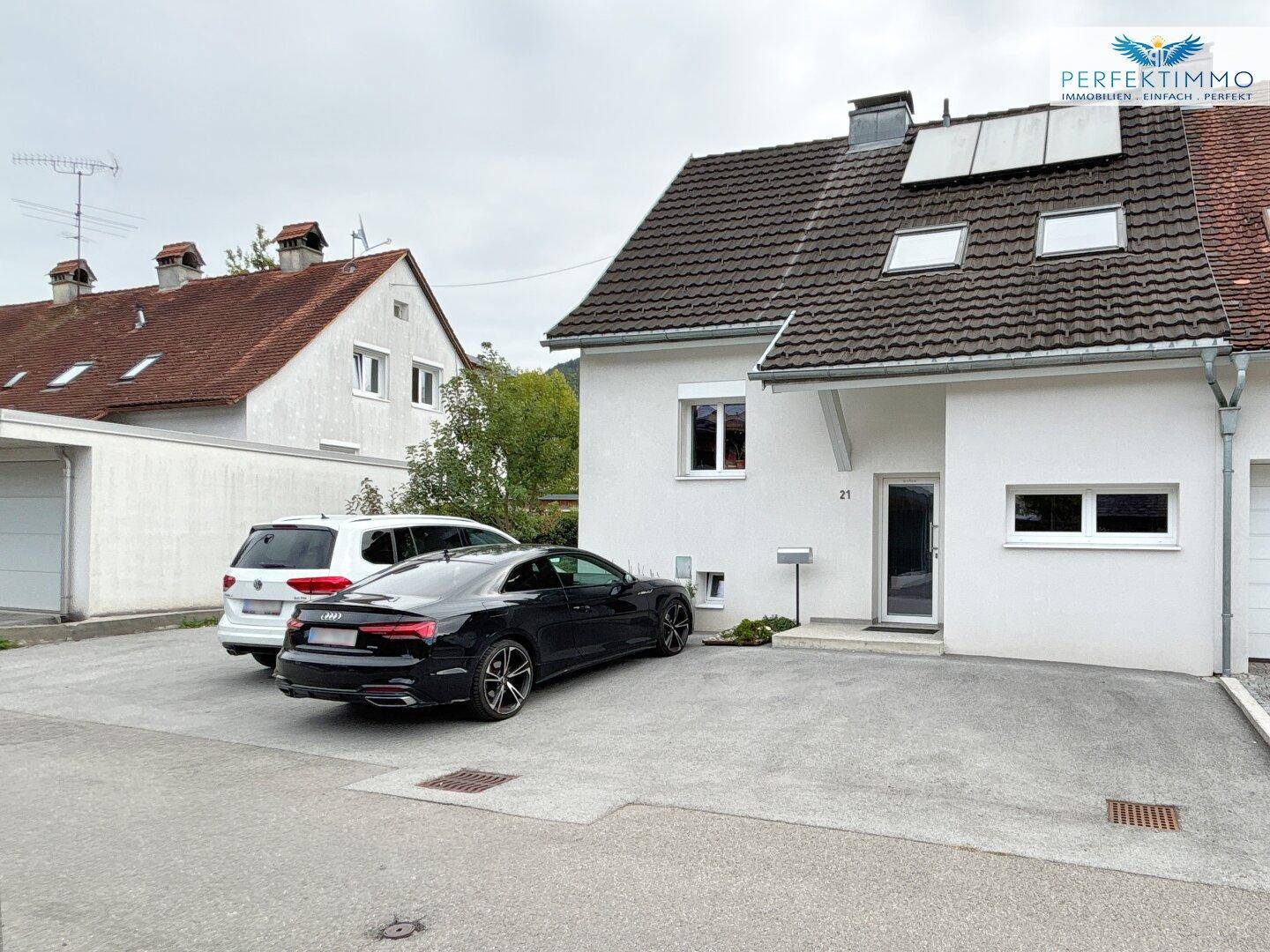 Vorderansicht des modernen Hauses mit weißer Fassade, Satteldach und Parkplätzen vor dem Eingang.