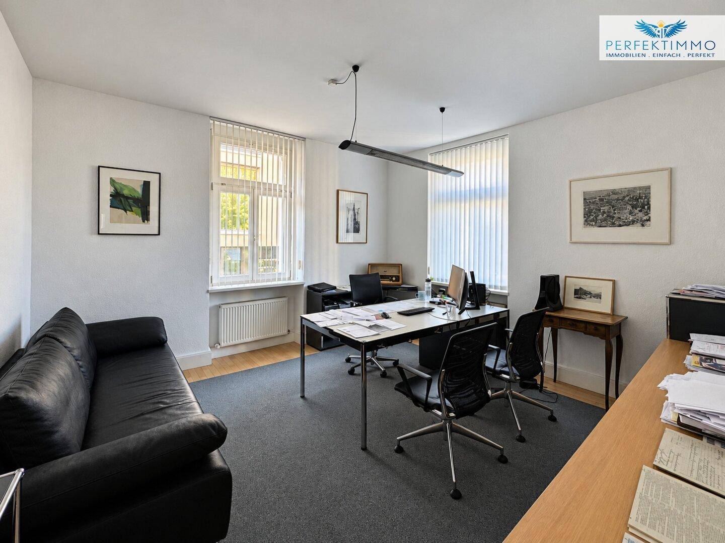 Helles Büro mit Teppichboden, großem Schreibtisch und schwarzem Sofa.