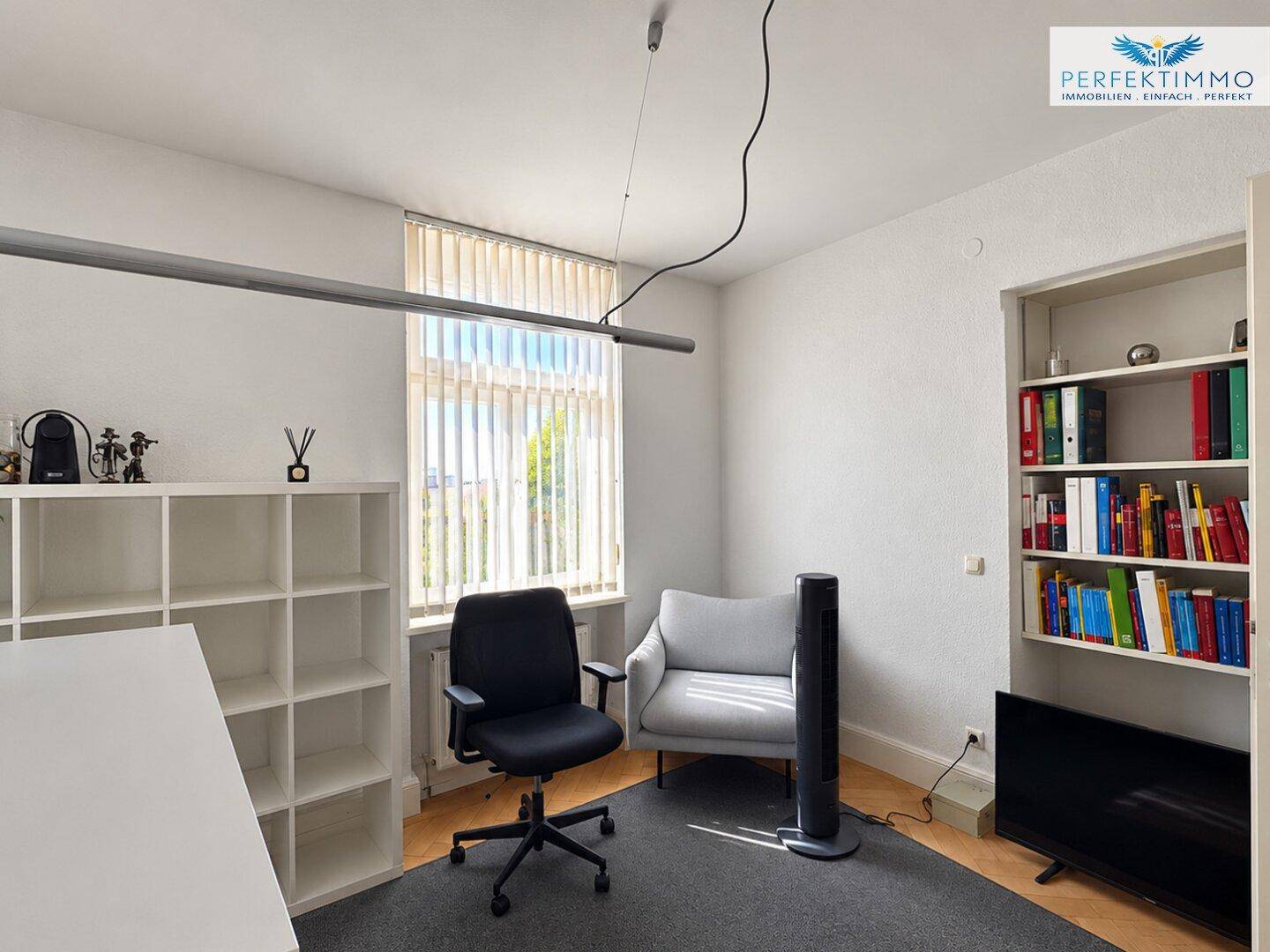 Ruhiges Büro mit Teppichboden, Bücherregal und einem Sessel am Fenster.