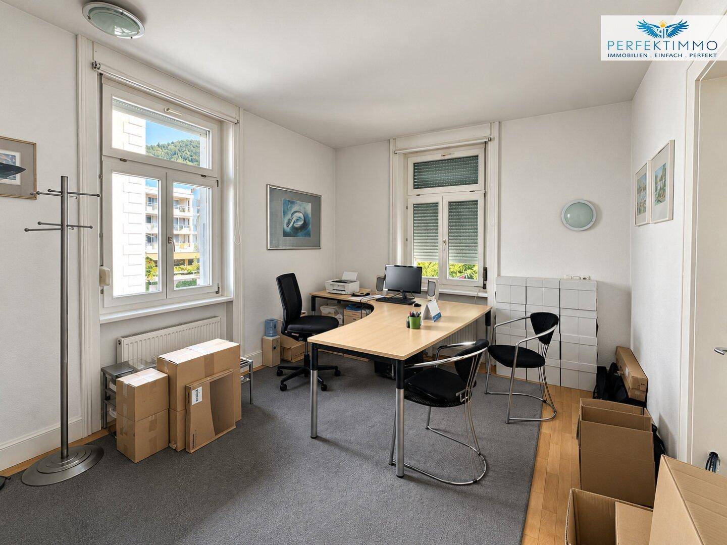 Helles Büro mit Teppichboden, zwei großen Fenstern und einem Schreibtisch.