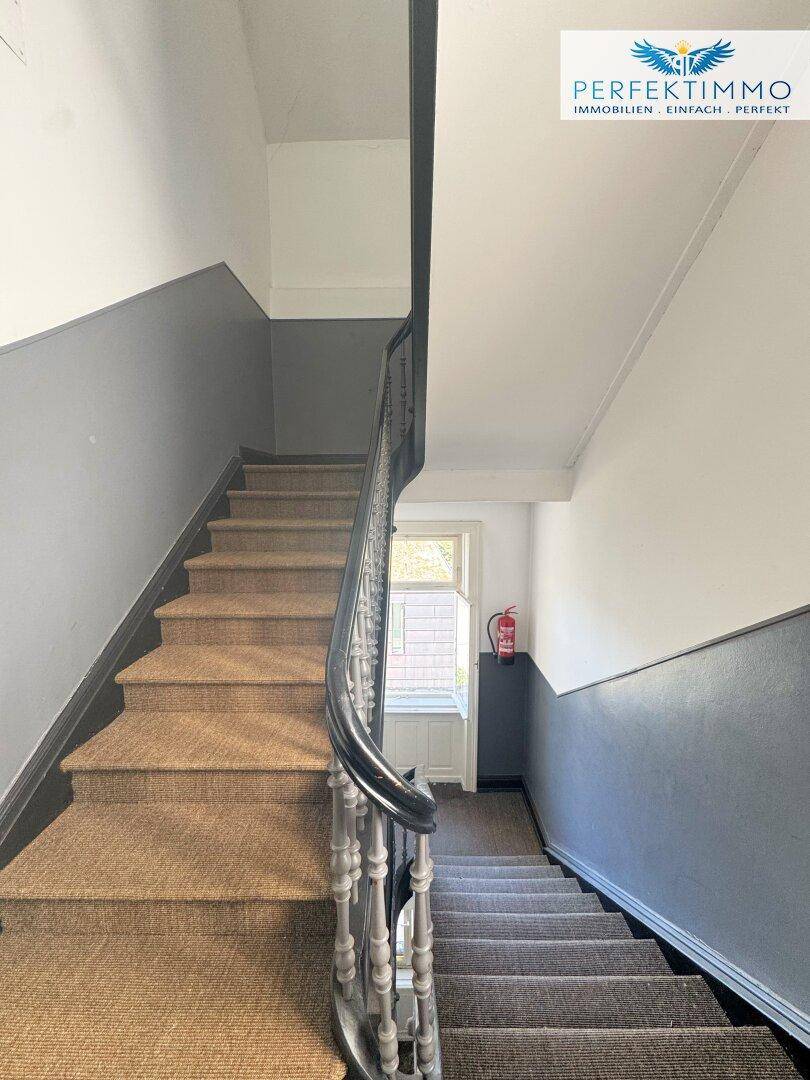 Treppenhaus mit Teppichstufen, dunklem Geländer und zweifarbigen Wänden.