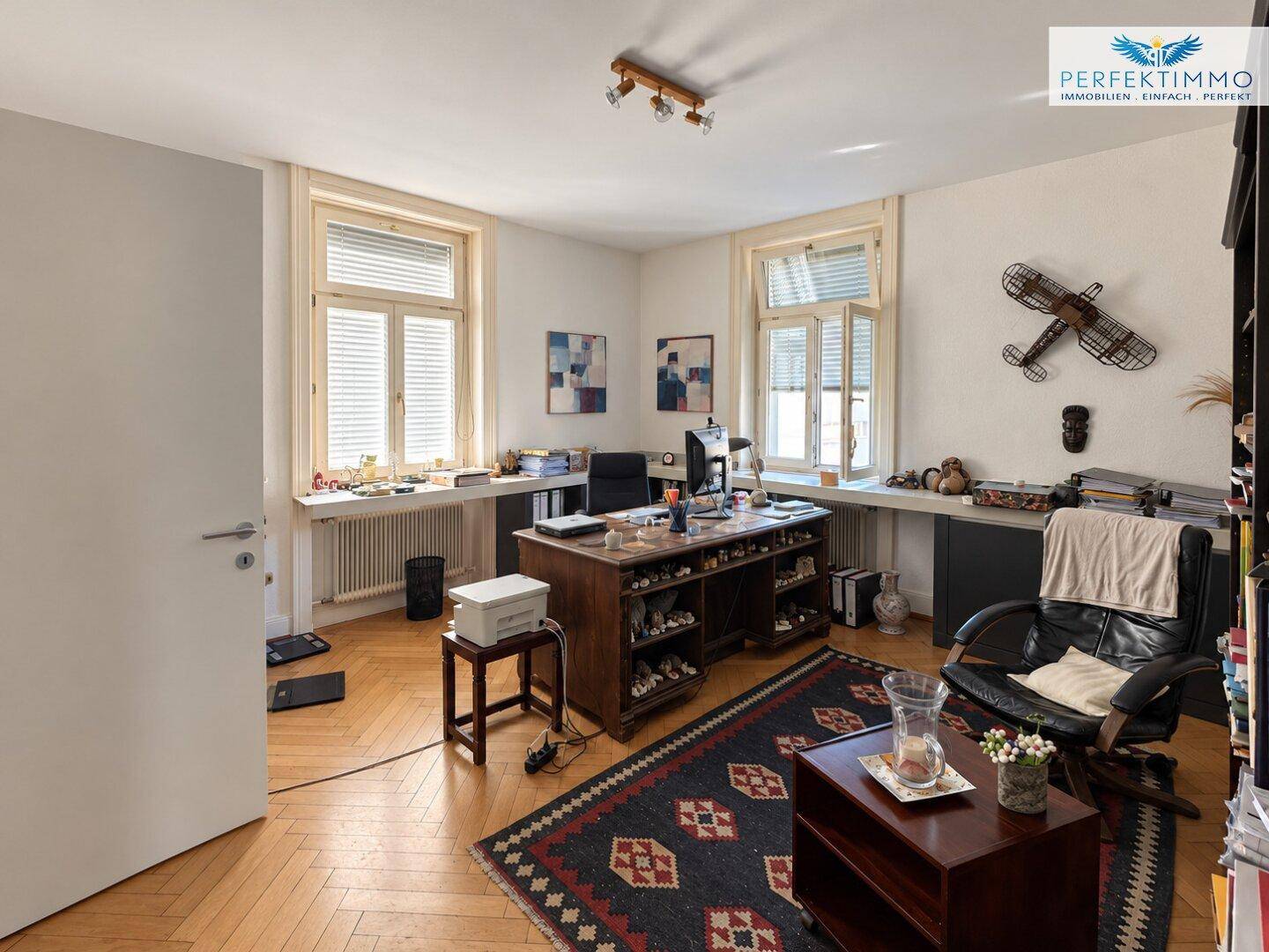 Geräumiges Büro mit Fischgrätparkett, großem Holzschreibtisch und Teppich.