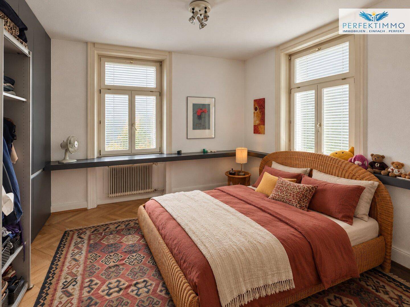 Schlafzimmer mit Rattanbett, Teppich und zwei großen Fenstern.