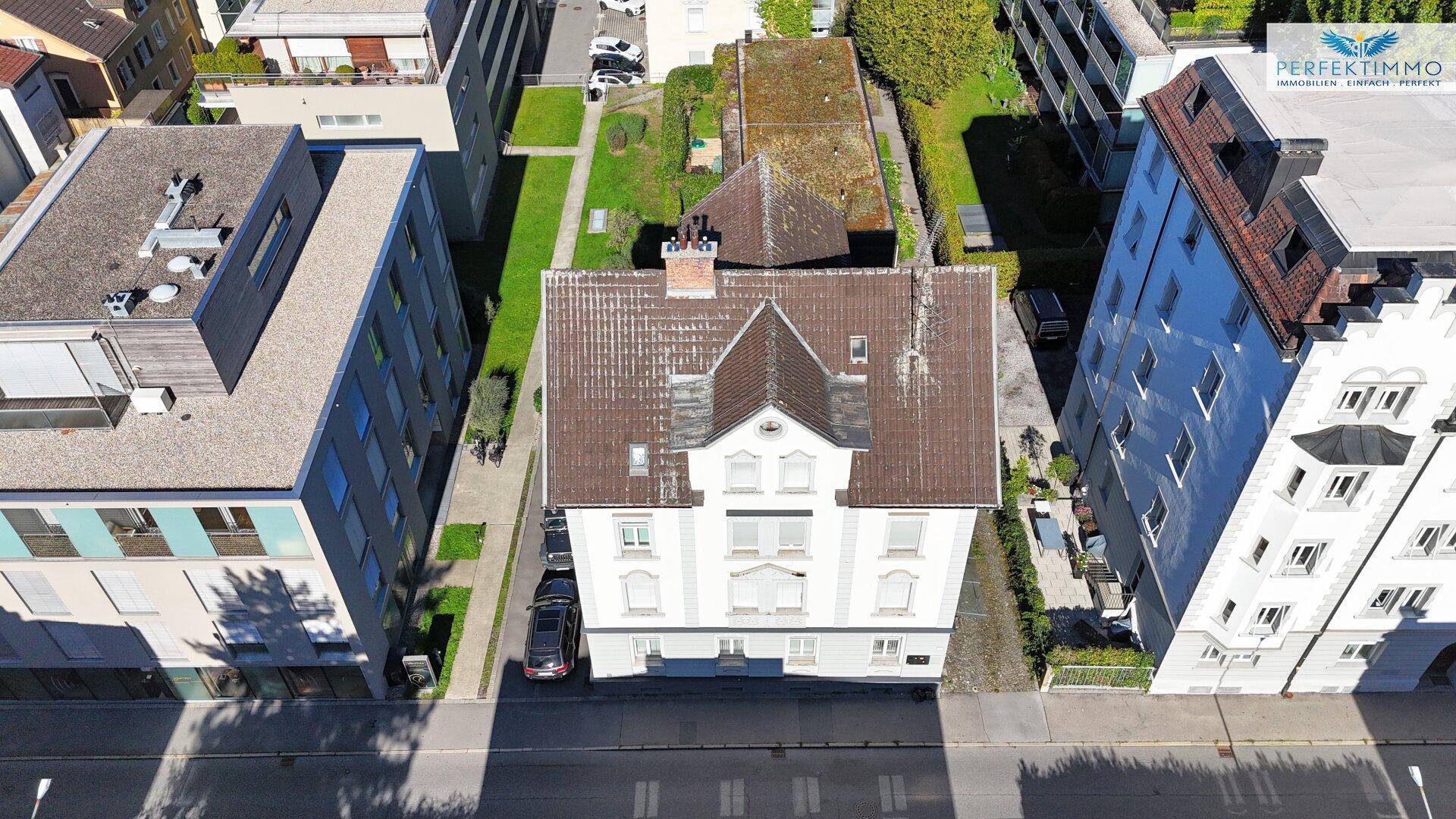 Luftaufnahme eines Mehrfamilienhauses mit Satteldach und umliegenden Gebäuden in städtischer Umgebung.