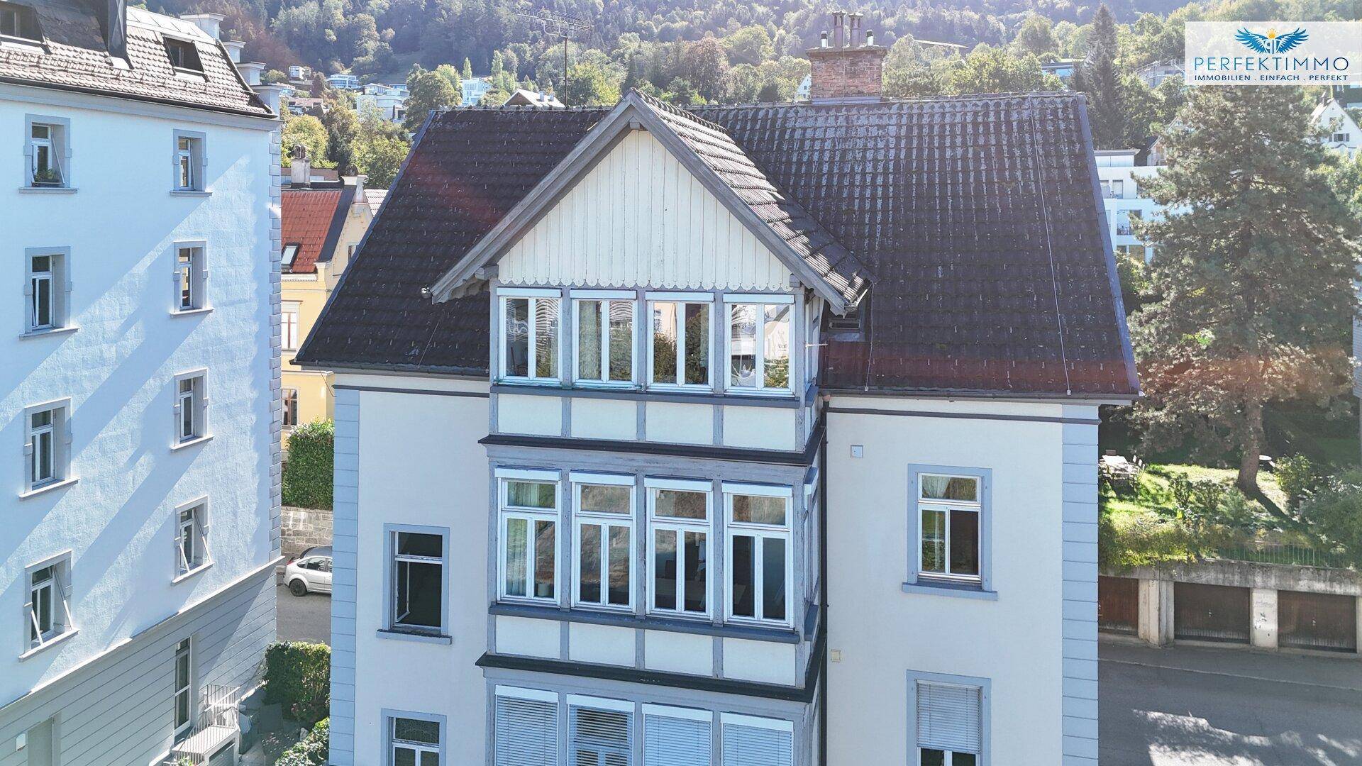 Fassade eines Altbaus mit markanten Erkern und vielen Fenstern, umgeben von grüner Landschaft.