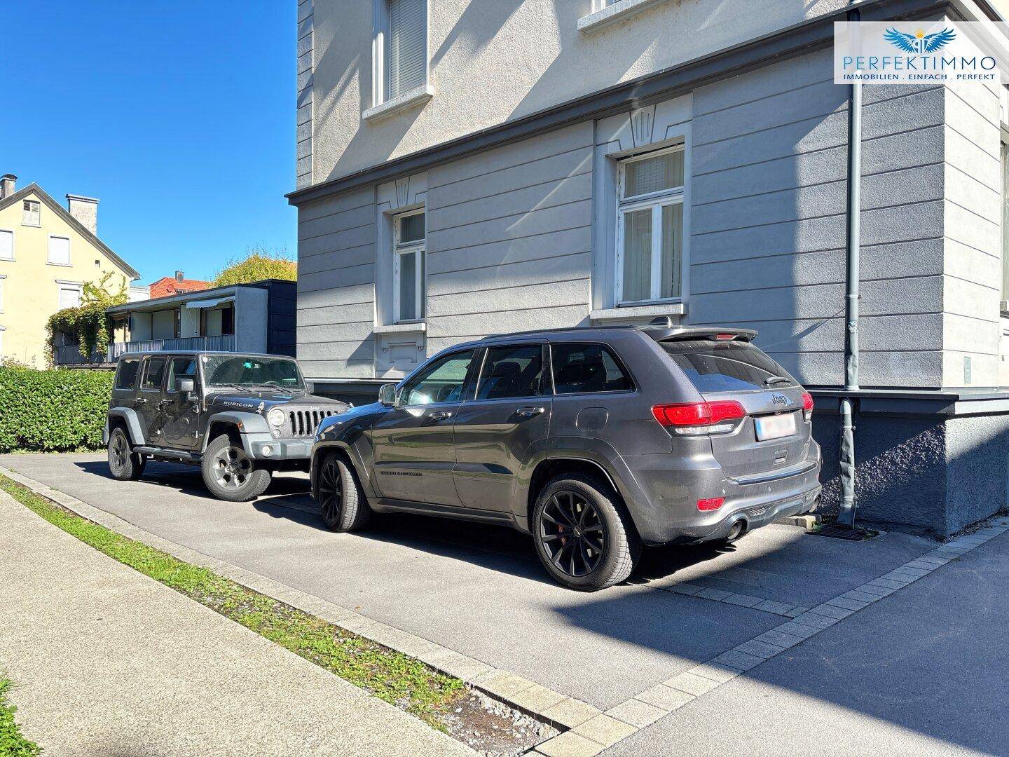Zwei Fahrzeuge parken auf einem Stellplatz vor der Fassade eines Wohngebäudes an einem sonnigen Tag.