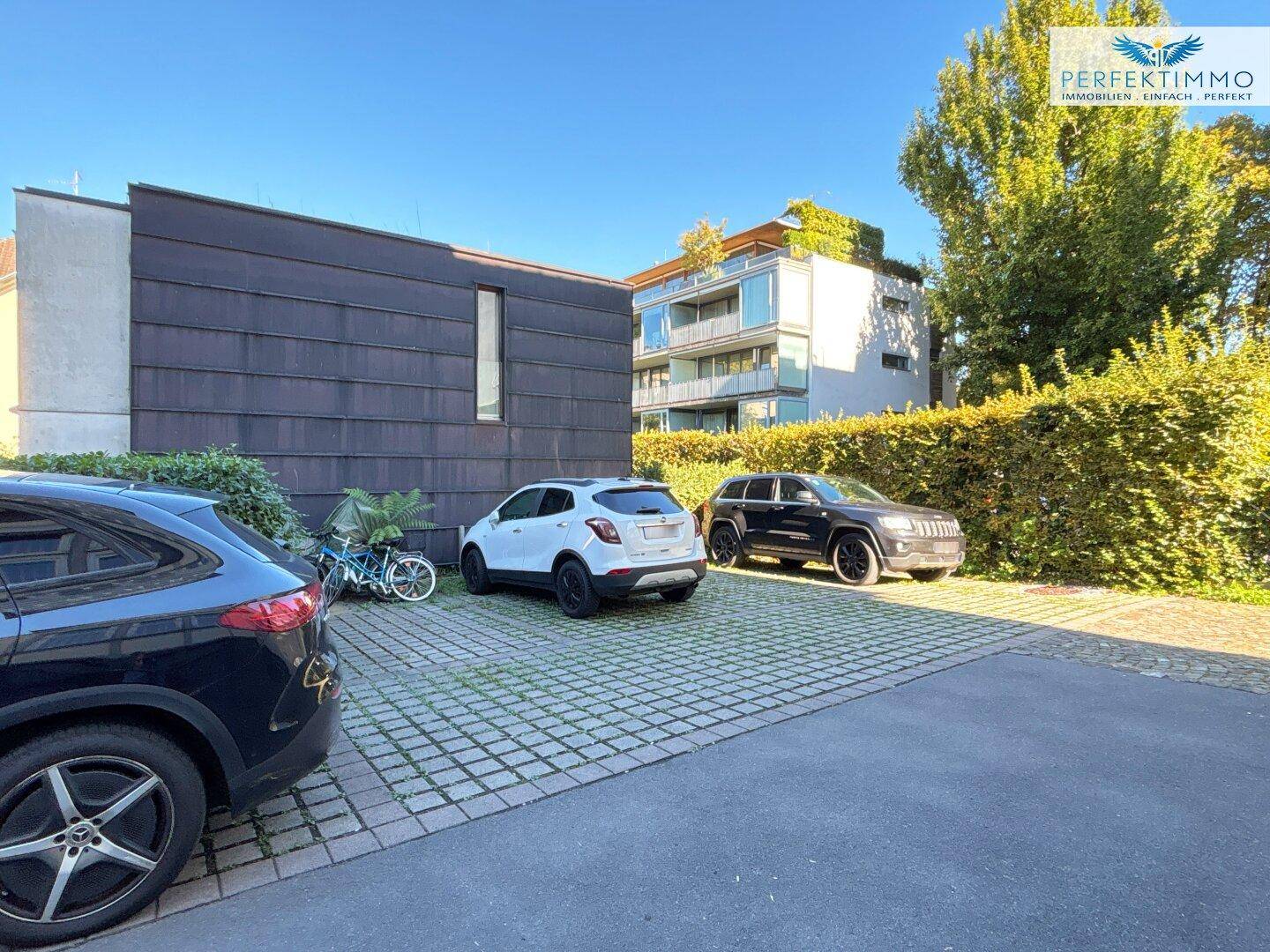 Mehrere Autos parken auf einem gepflasterten Bereich, umgeben von einer hohen Hecke und Bäumen.