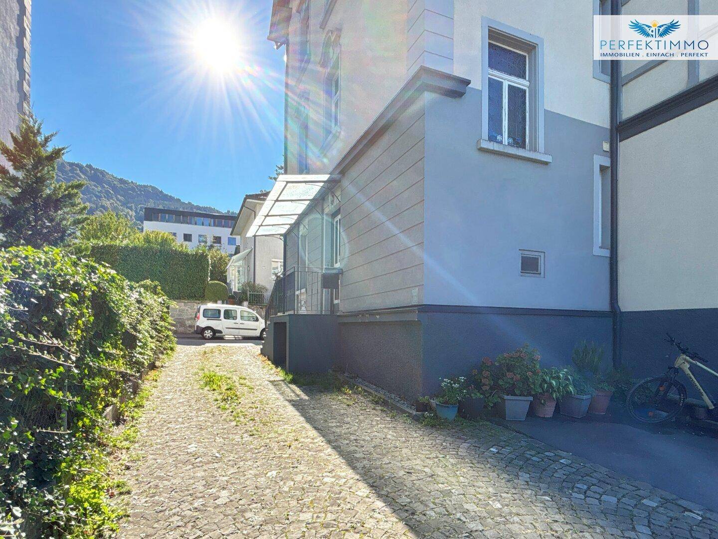 Sonnenbeschienene Auffahrt neben einem Gebäude mit üppiger Vegetation und Blick auf die Berge im Hintergrund.
