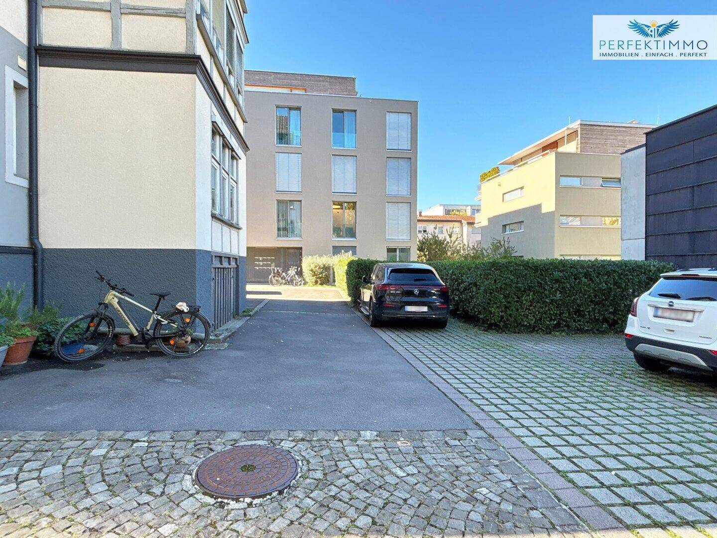 Gepflasterter Parkplatz mit Fahrrädern und Autos, umgeben von Hecken und modernen Gebäuden.