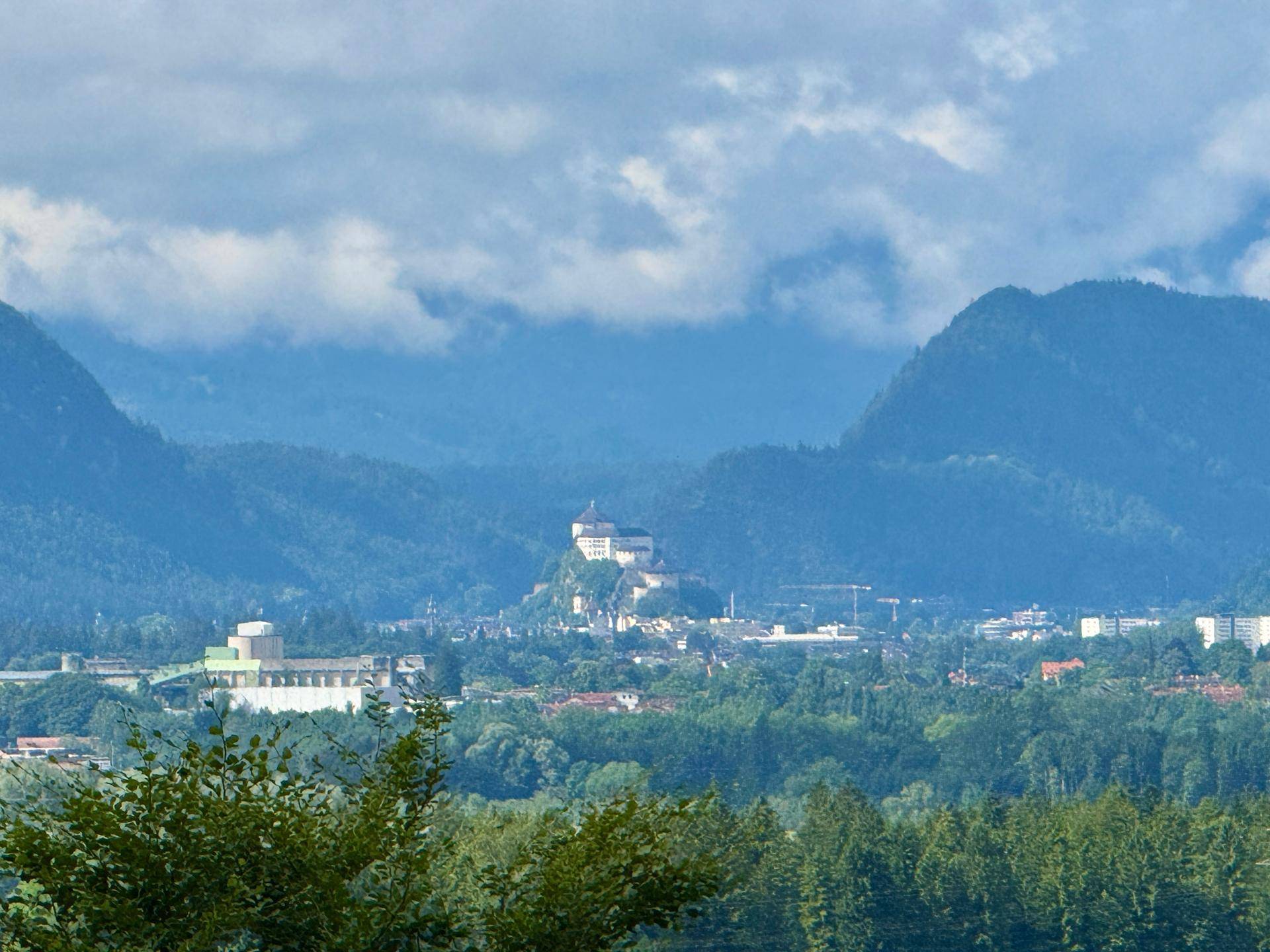 Panoramablick auf eine Stadt mit einer markanten Burg, umgeben von Bergen unter bewölktem Himmel.