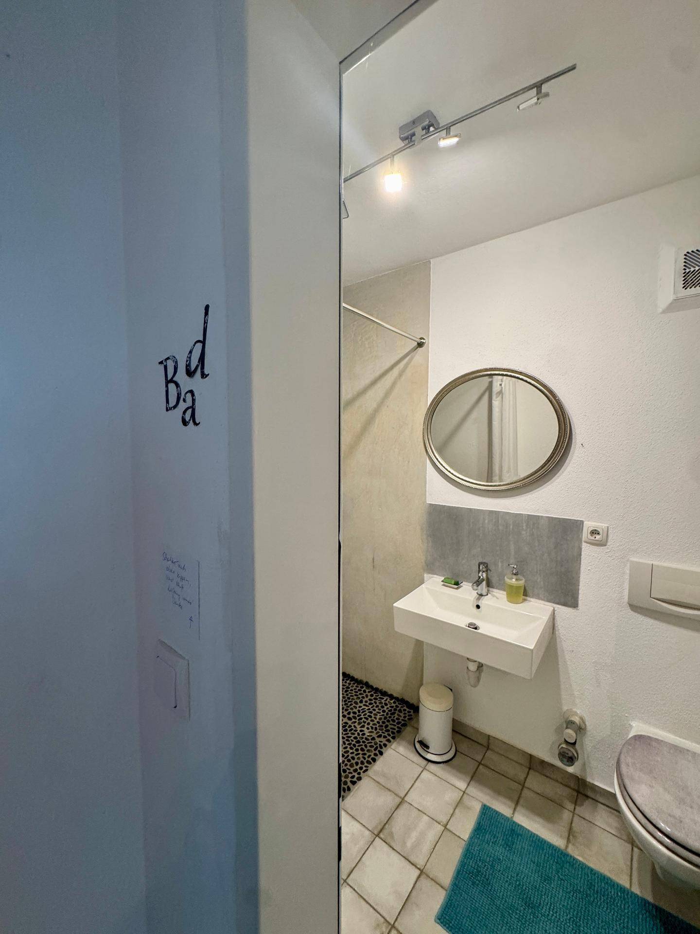 Modernes Badezimmer mit wandhängendem Waschbecken, ovalem Spiegel und Duschbereich.