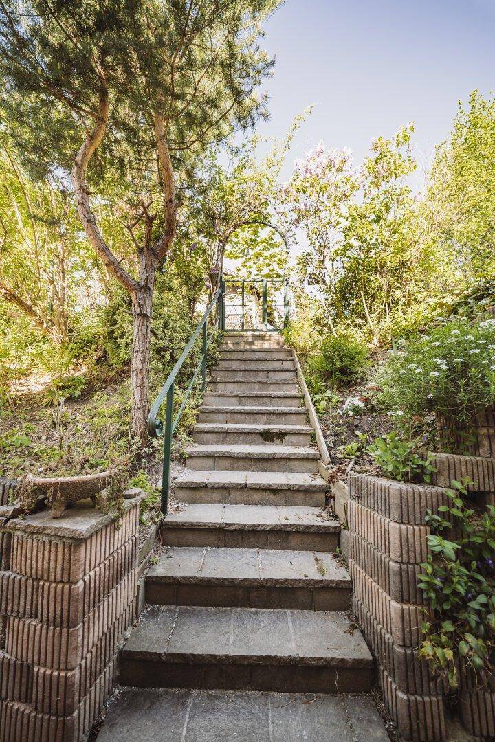 Lange Steintreppe mit Handlauf, umgeben von üppiger Vegetation im Garten.