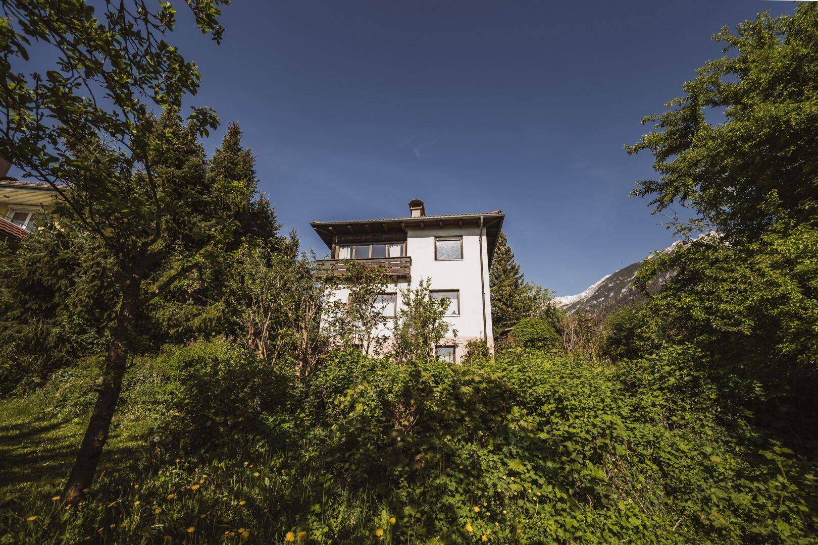 Hausfassade mit Balkon, umgeben von Bäumen und Sträuchern, mit Bergblick.