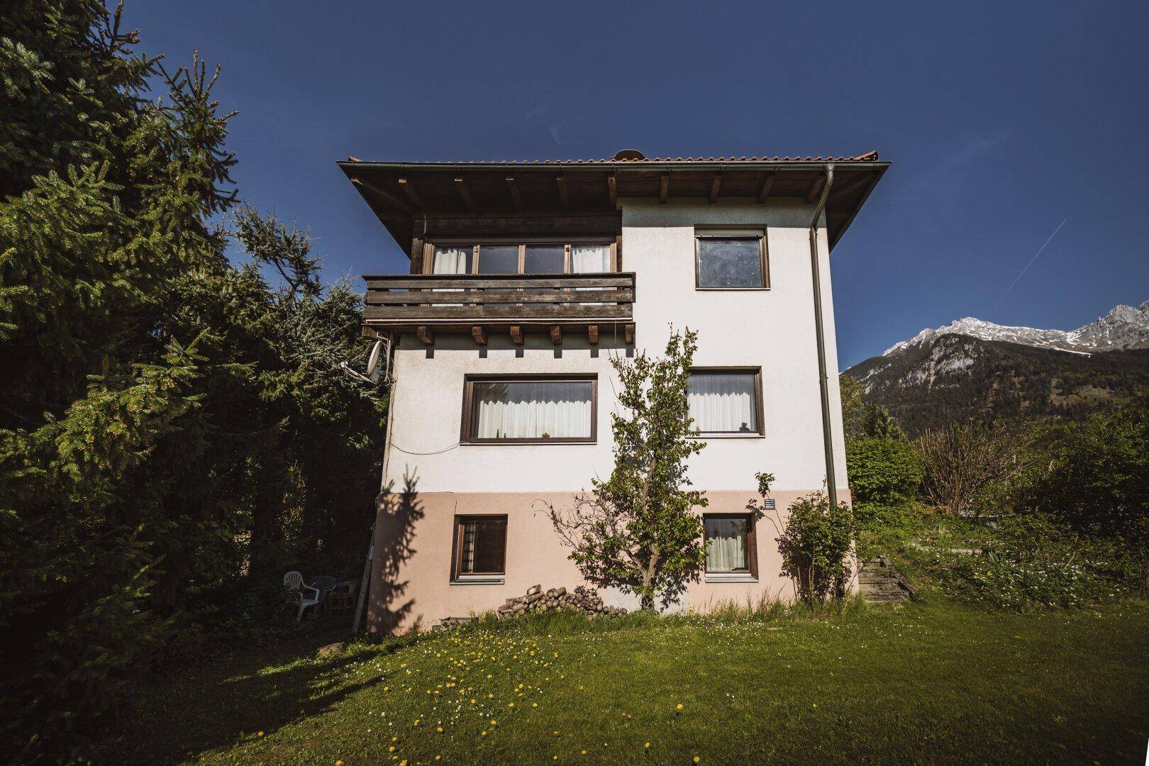 Frontansicht des Hauses mit Balkon, umgeben von grüner Wiese und Bäumen.