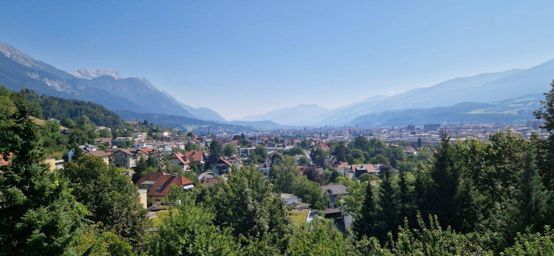 Weitläufiger Blick über eine Stadt im Tal, umgeben von Bergen und viel Grün.