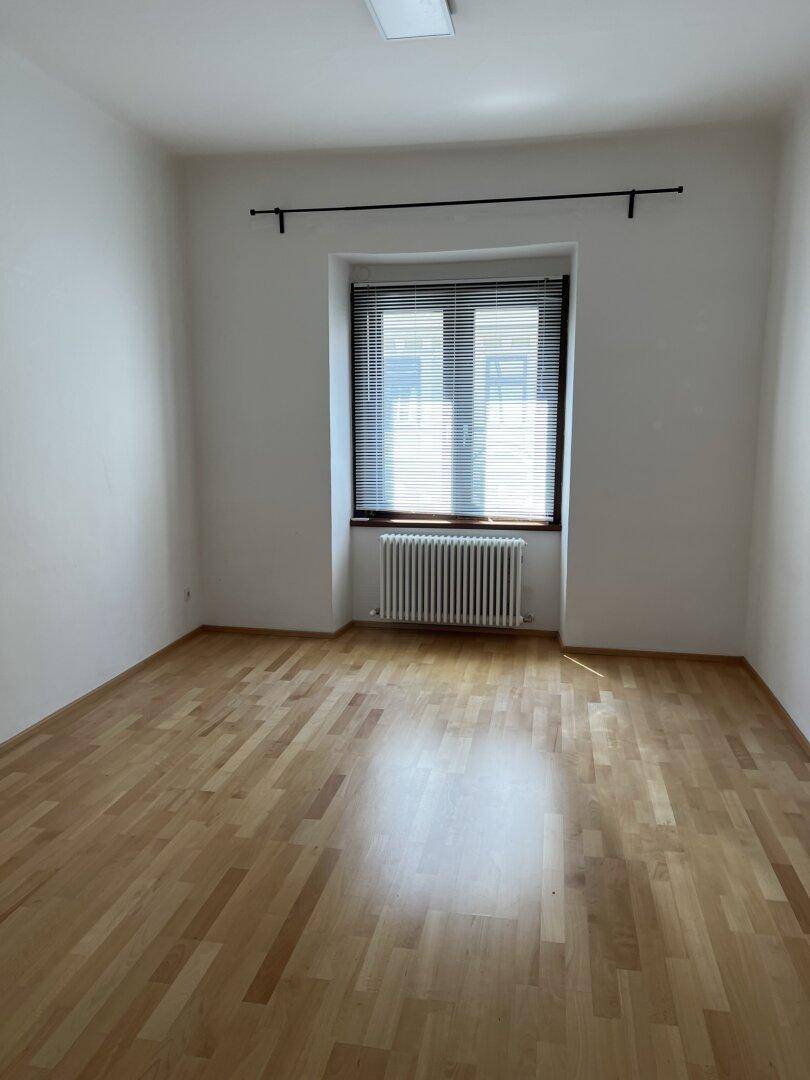 Helles Schlafzimmer mit Parkettboden, Fenster mit Jalousien und Heizkörper unter dem Fenster.