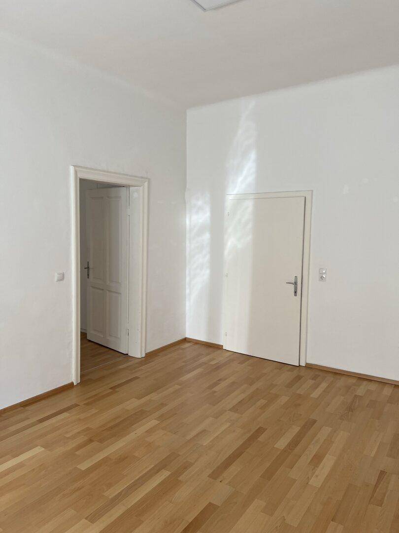 Leeres Zimmer mit hellem Parkettboden und zwei weißen Türen, die zu anderen Räumen führen.