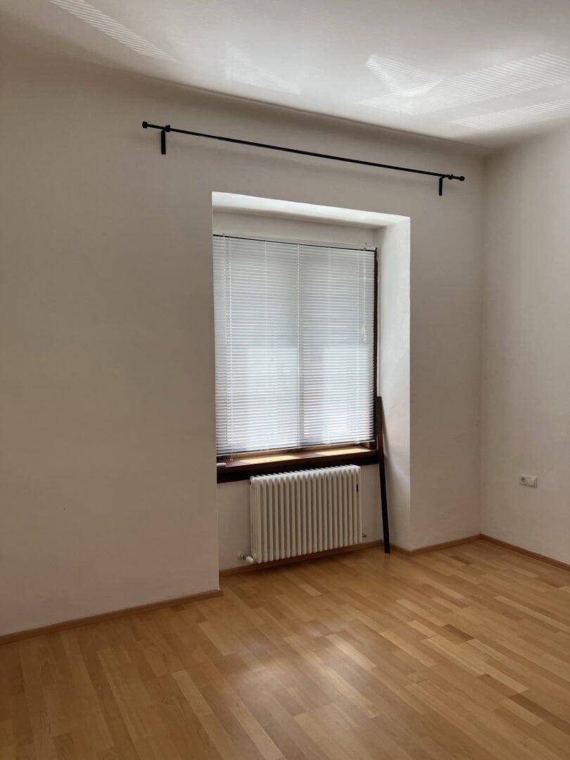 Leeres Zimmer mit Parkettboden, großem Fenster mit Jalousien und Heizung.