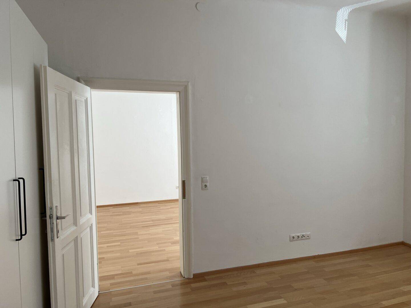 Helles Zimmer mit Parkettboden und weißer Tür, die in einen weiteren Raum führt.