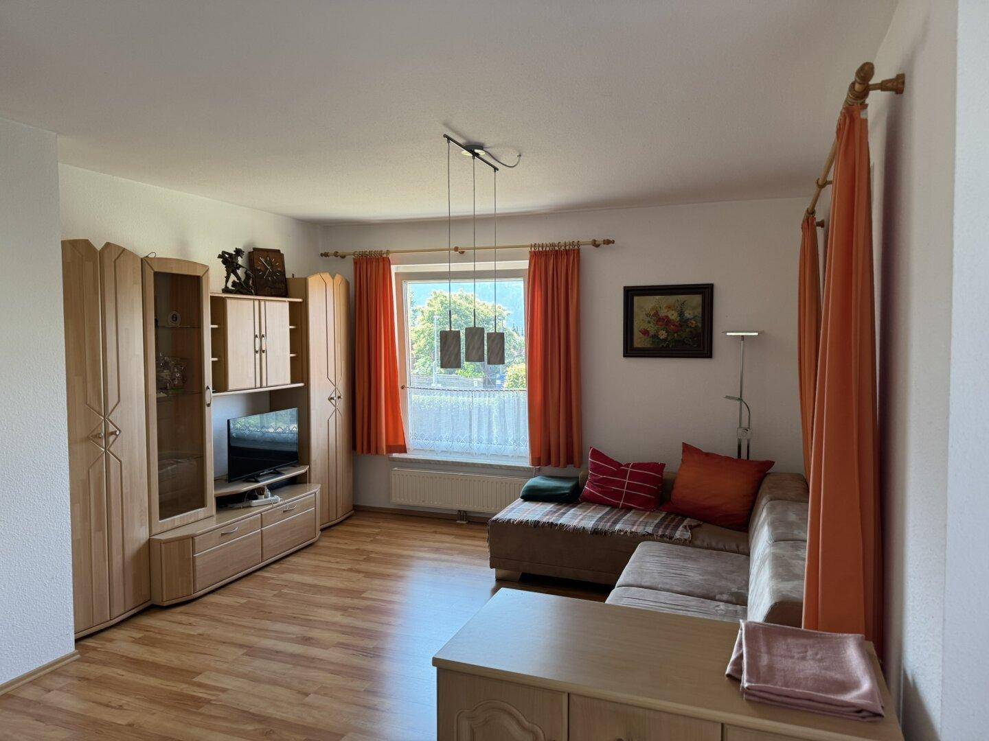 Wohnzimmer mit Sofa, Laminatboden und Fenster mit orangefarbenen Vorhängen.