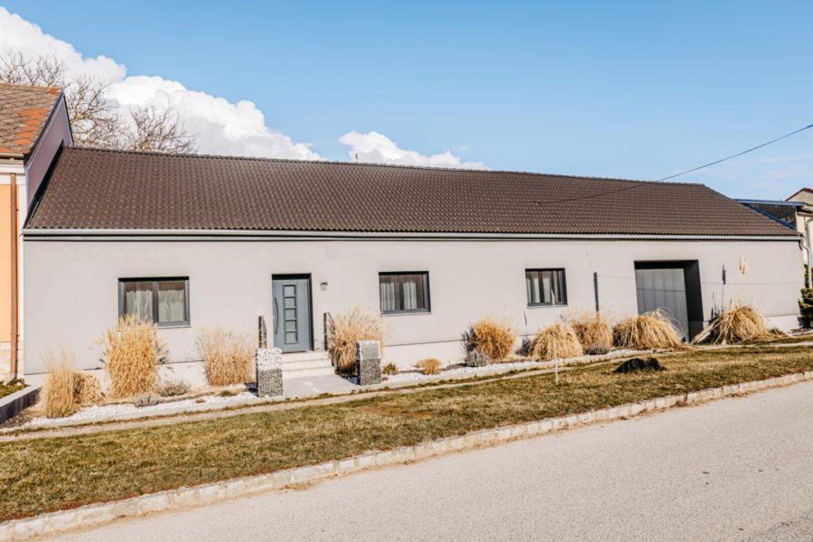 Ansicht des modernen Hauses mit heller Fassade, dunklem Dach und gepflegter Außenanlage an einem sonnigen Tag.
