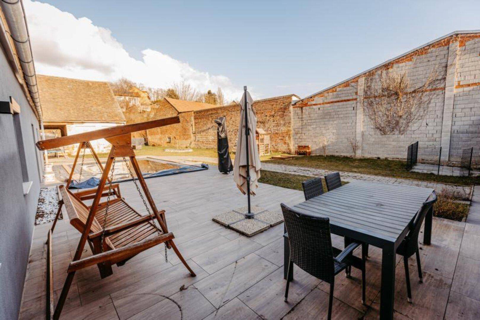 Geräumige Terrasse mit Sitzgelegenheiten, Schaukelbank und einem Pool im Hintergrund des Gartens.