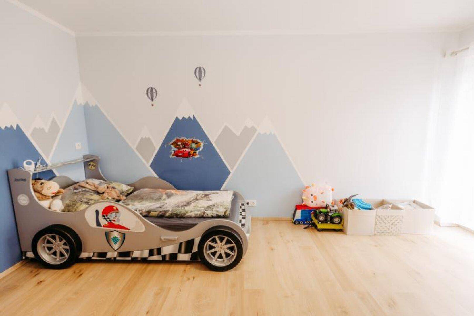 Kreativ gestaltetes Kinderzimmer mit Rennwagenbett, Holzboden und Berglandschaft an der Wand.