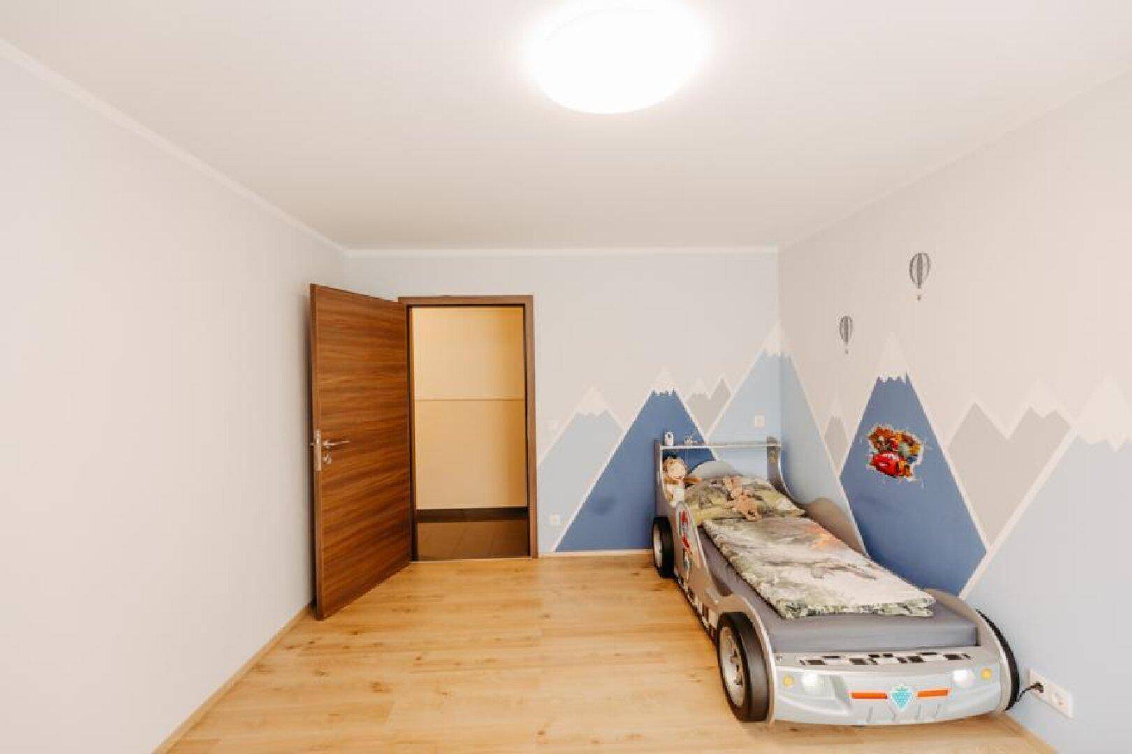 Kinderzimmer mit Rennwagenbett, hellem Holzboden und Bergmotiv-Wandmalerei, bereit zum Spielen.