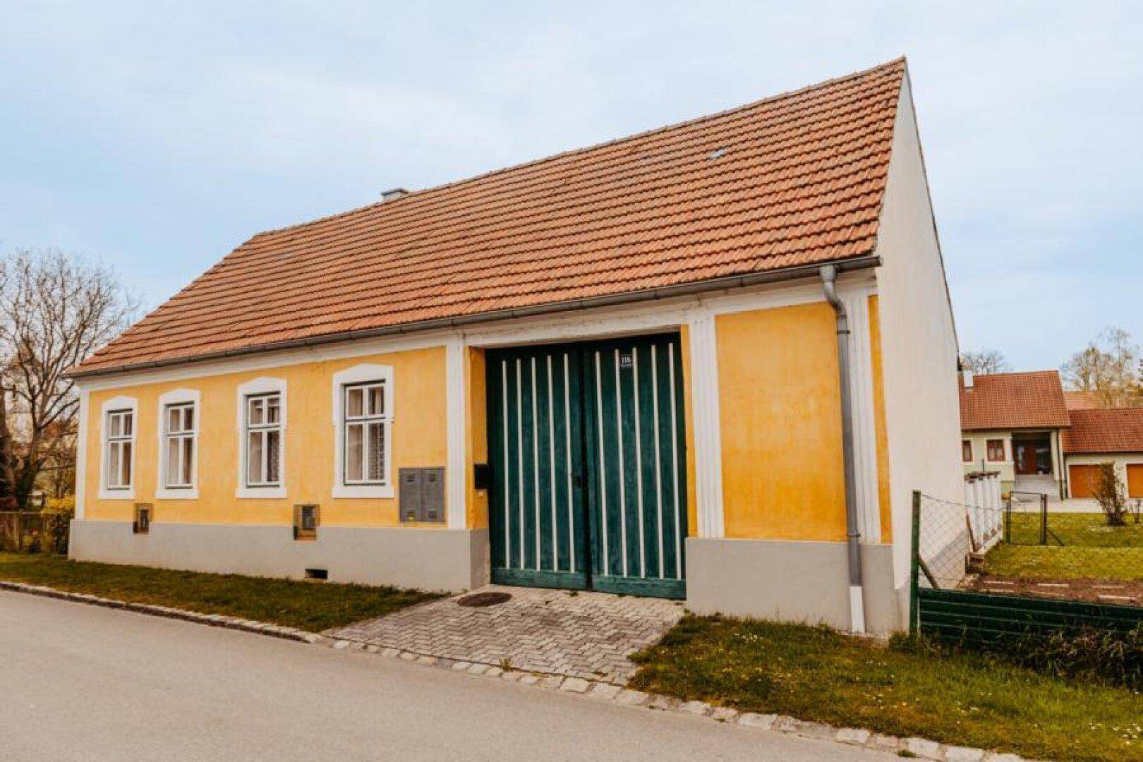 Gelbes Landhaus mit rotem Ziegeldach und großen grünen Scheunentoren von der Seite.
