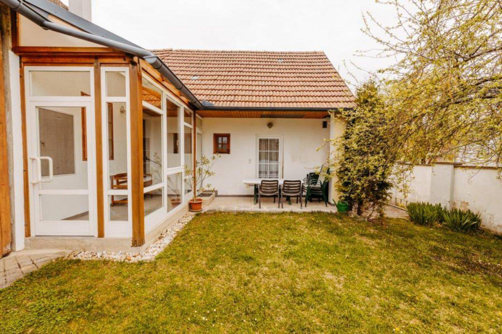 Garten mit Rasenfläche, angrenzendem Wintergarten und kleiner Terrasse mit Sitzgelegenheiten.