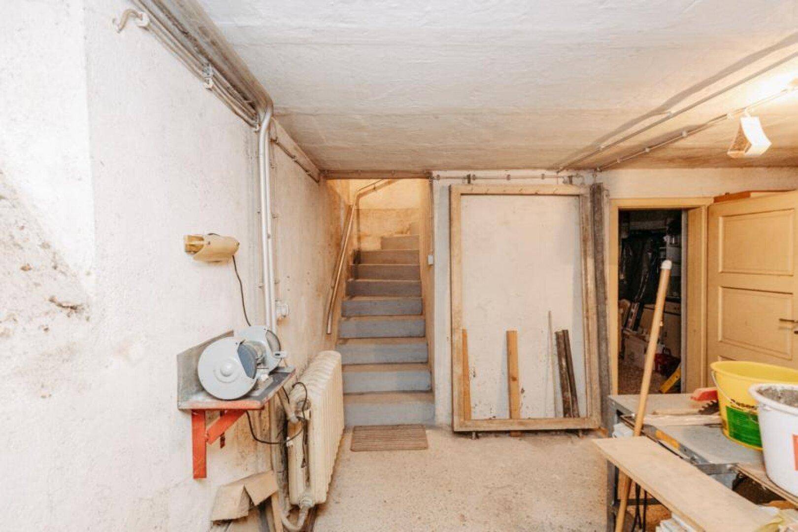 Kellerraum mit Treppenaufgang, Heizkörper und Werkbank im Hintergrund.