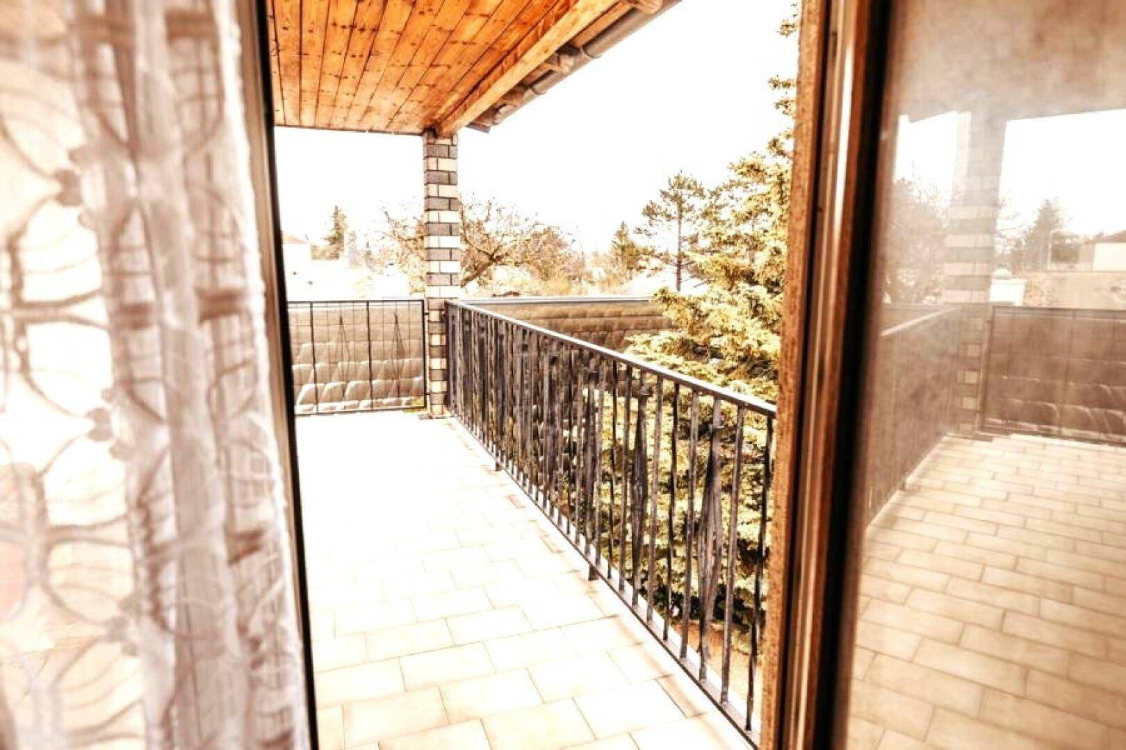 Überdachter Balkon mit Fliesenboden und Metallgeländer, umgeben von Bäumen.