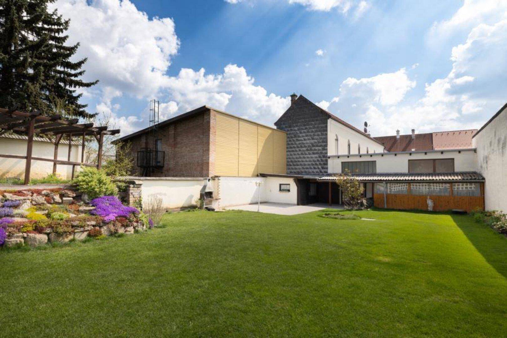 Gepflegter Gartenbereich mit Rasenfläche, Blumenbeeten und alten Bäumen hinter dem Gebäude.