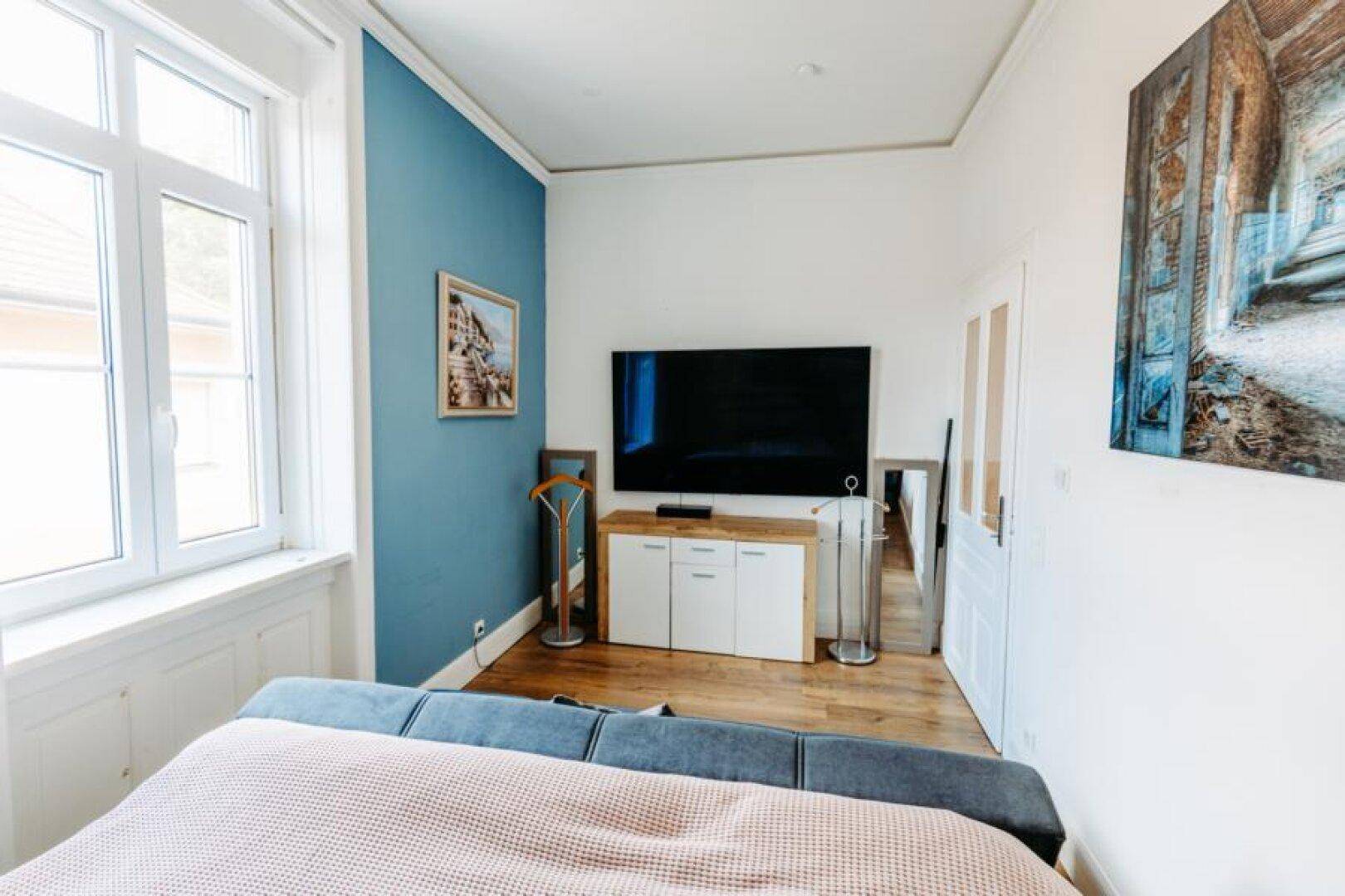 Schlafzimmer mit großem Fernseher, Fenster und blauer Akzentwand.
