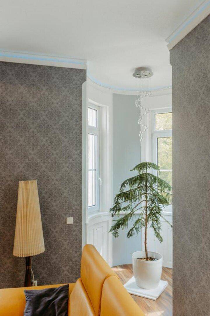 Gemütliches Wohnzimmer mit gelbem Sofa, gemusterter Tapete und Erkerfenster mit Pflanze.