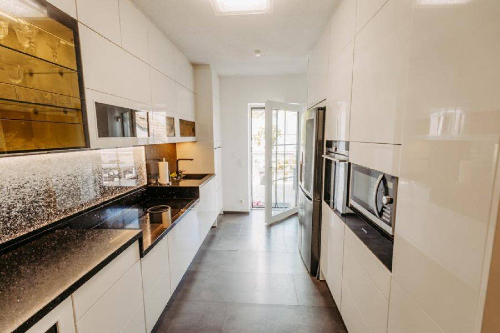 Moderne weiße Küche mit schwarzen Arbeitsplatten, Einbaugeräten und Zugang nach draußen.