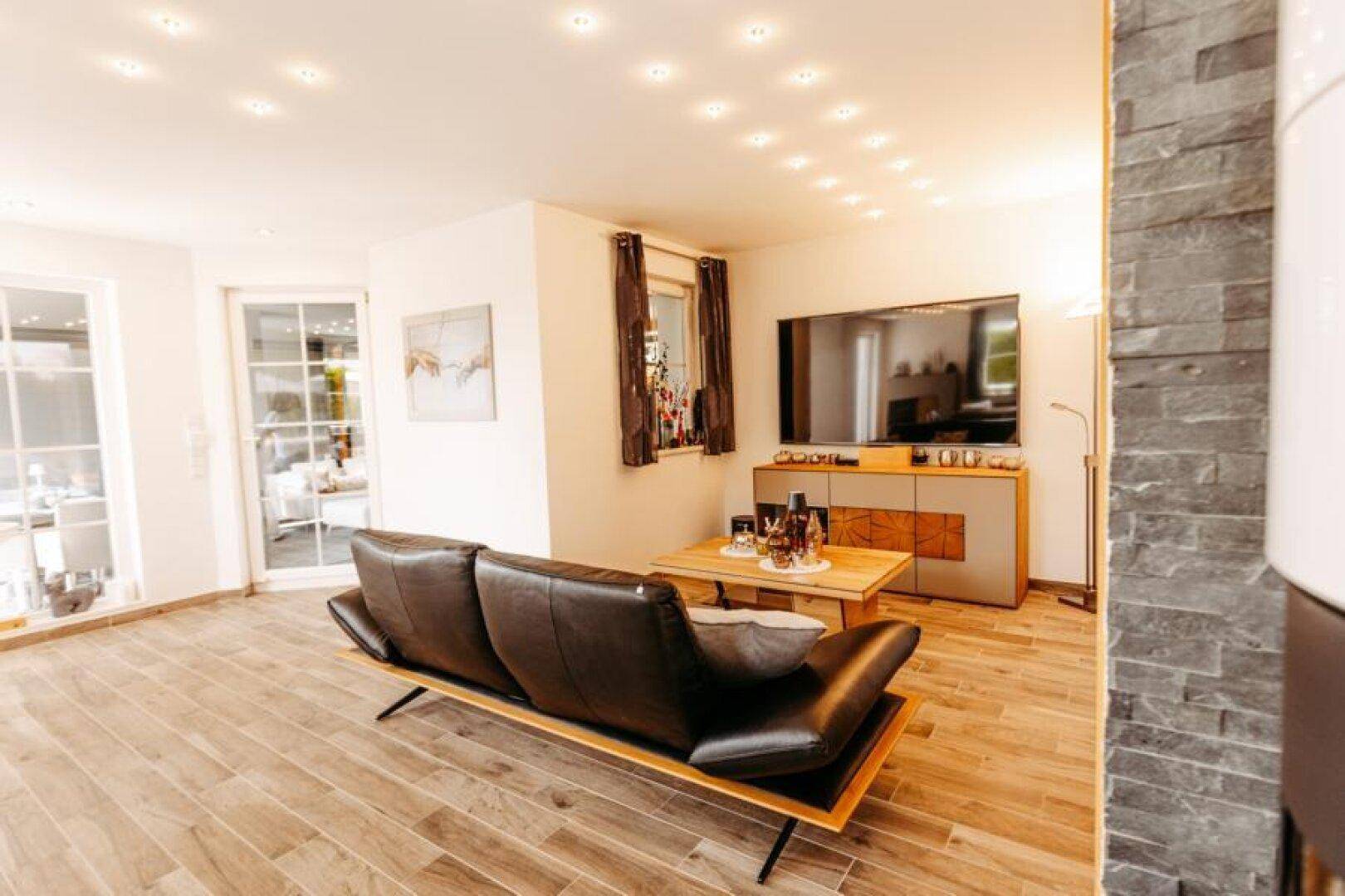 Gemütliches Wohnzimmer mit Ledersofa, großem Fernseher und einem modernen Kaminofen an der Steinwand.