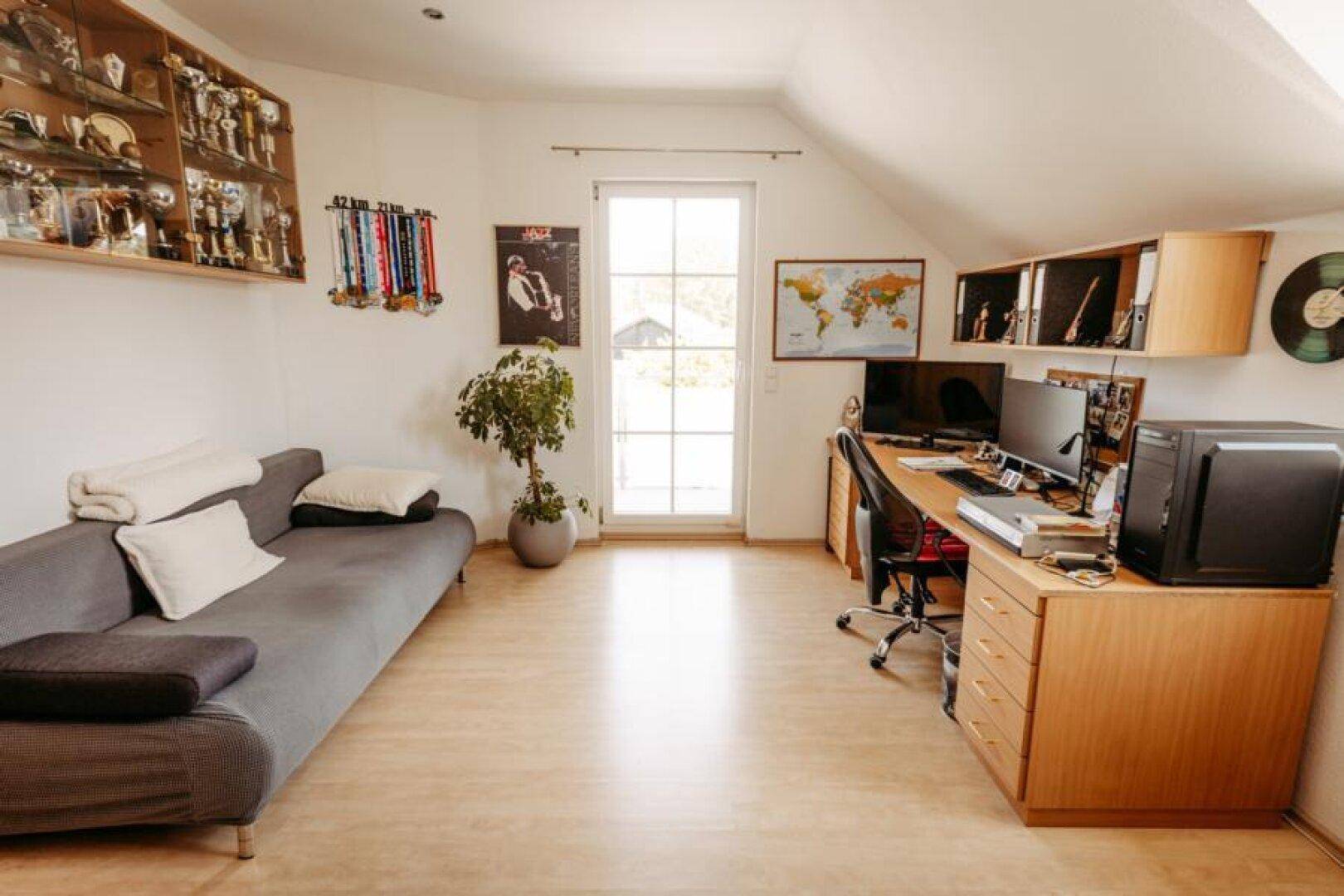 Helles Arbeitszimmer mit Schreibtisch, Sofa und Zugang zu einem Außenbereich, unter einem Dachschrägenfenster.