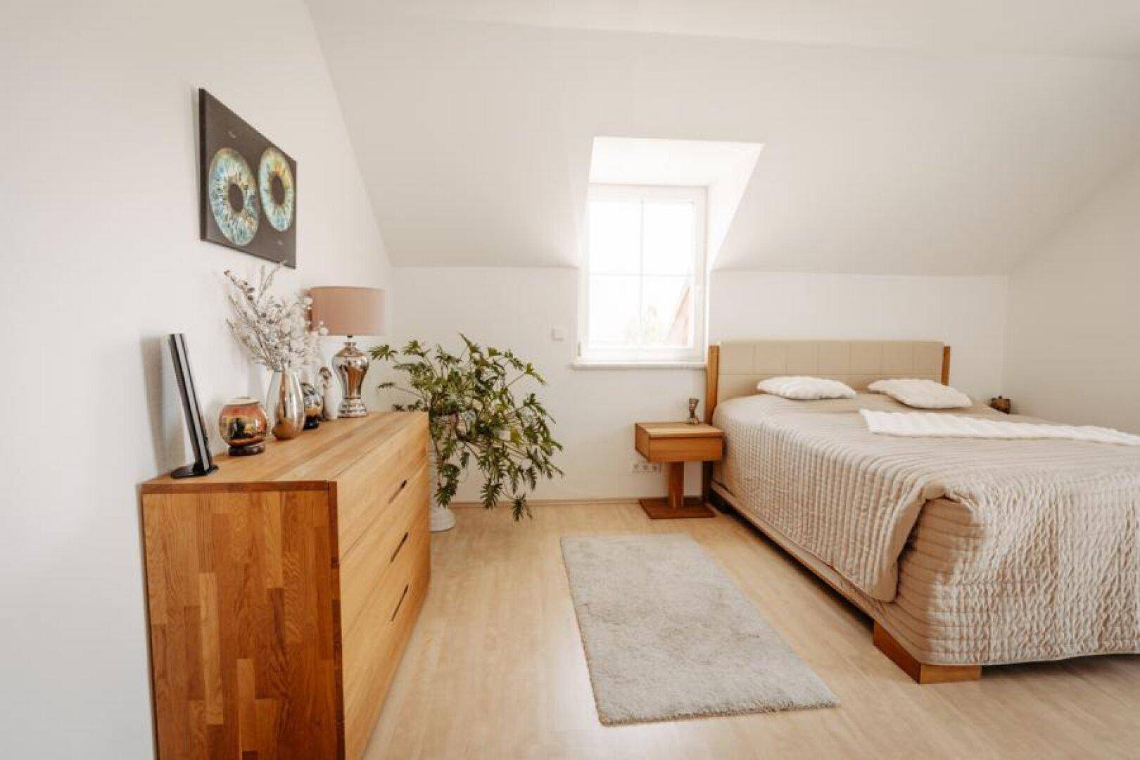 Helles Schlafzimmer mit Holzboden, Kommode und einem Fenster in der Dachschräge.