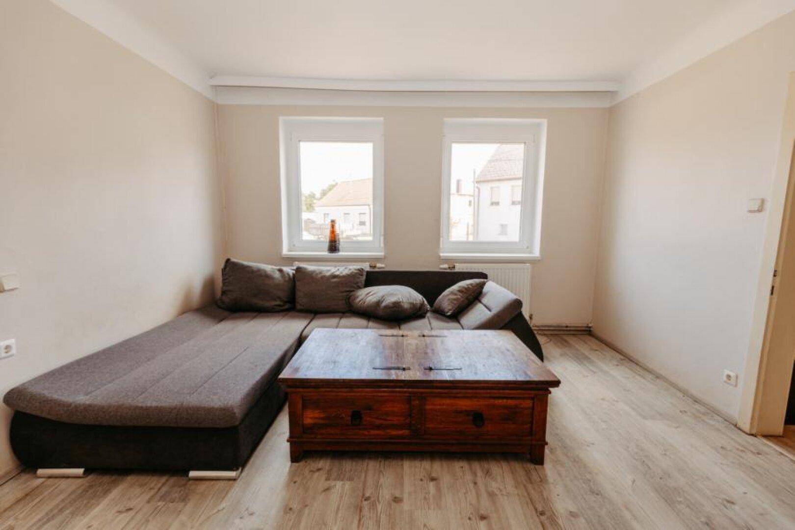 Gemütliches Wohnzimmer mit Ecksofa, Holztisch und Laminatboden, hell durch zwei Fenster.