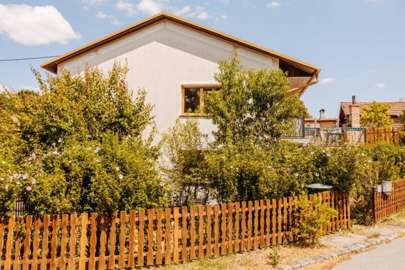 Seitenansicht eines Hauses mit Holzzaun und üppiger Bepflanzung unter sonnigem Himmel.