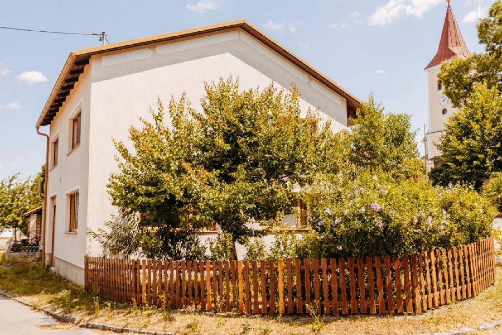 Seitenansicht des Hauses mit Holzzaun, Bäumen und einem Kirchturm im Hintergrund.