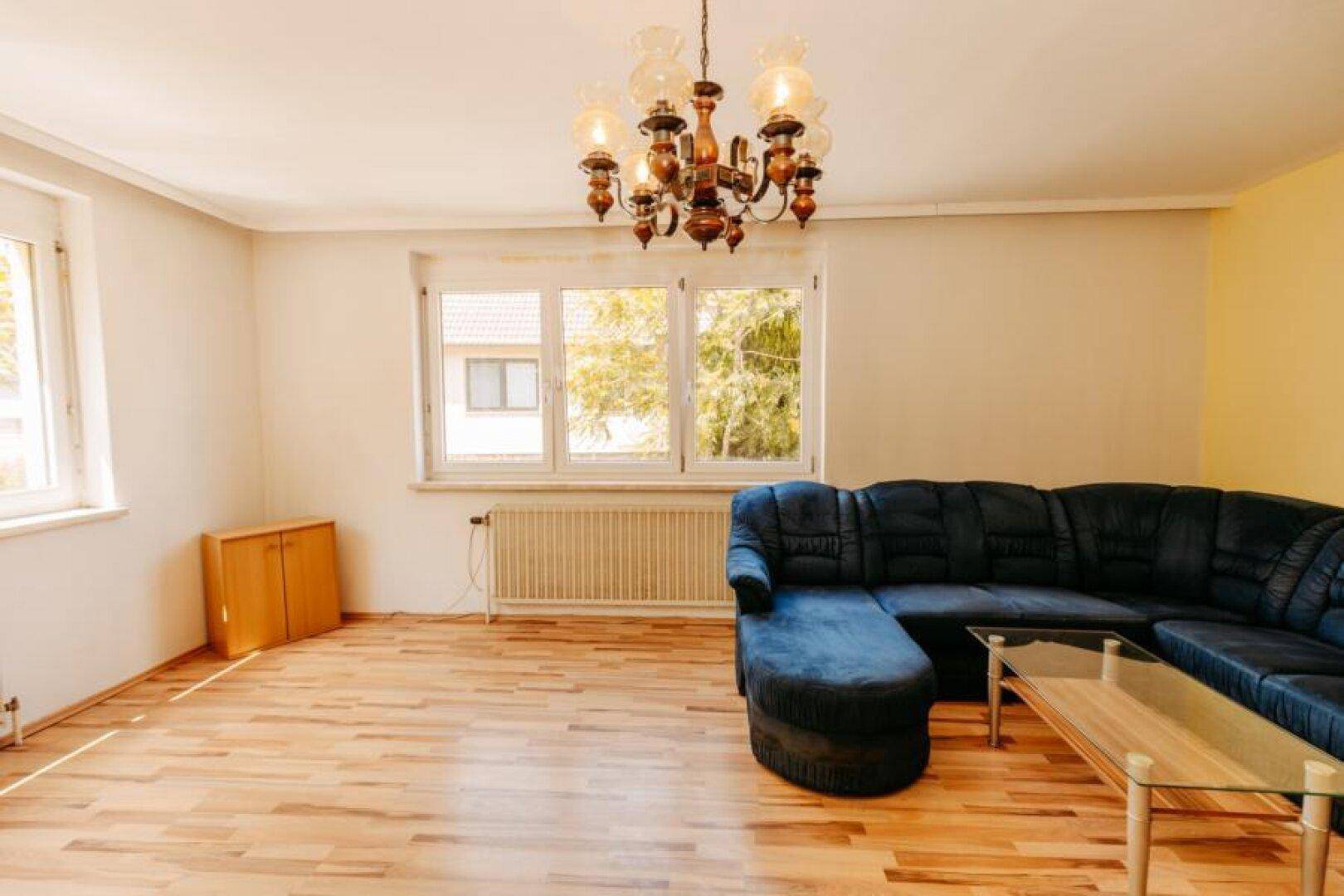 Geräumiges Wohnzimmer mit Parkettboden, großem Fenster und einem blauen Sofa.