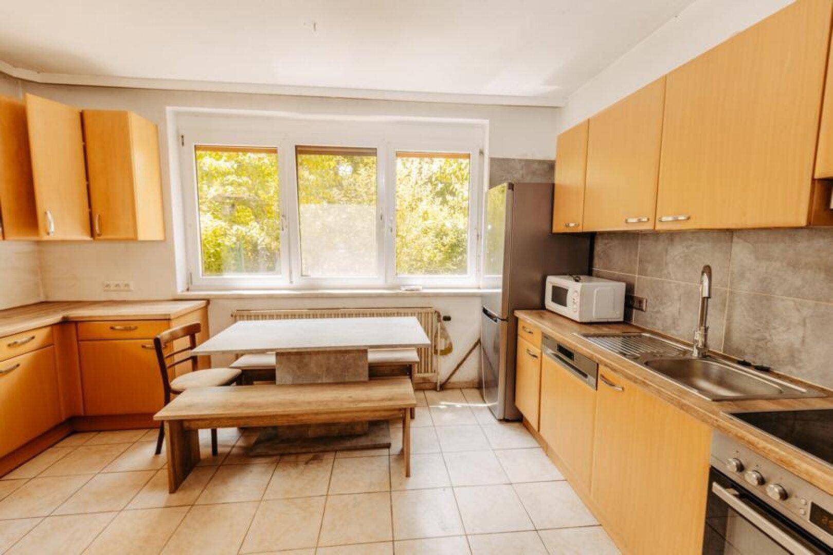 Helle Küche mit Holzschränken, Essbereich und Blick ins Grüne durch große Fenster.