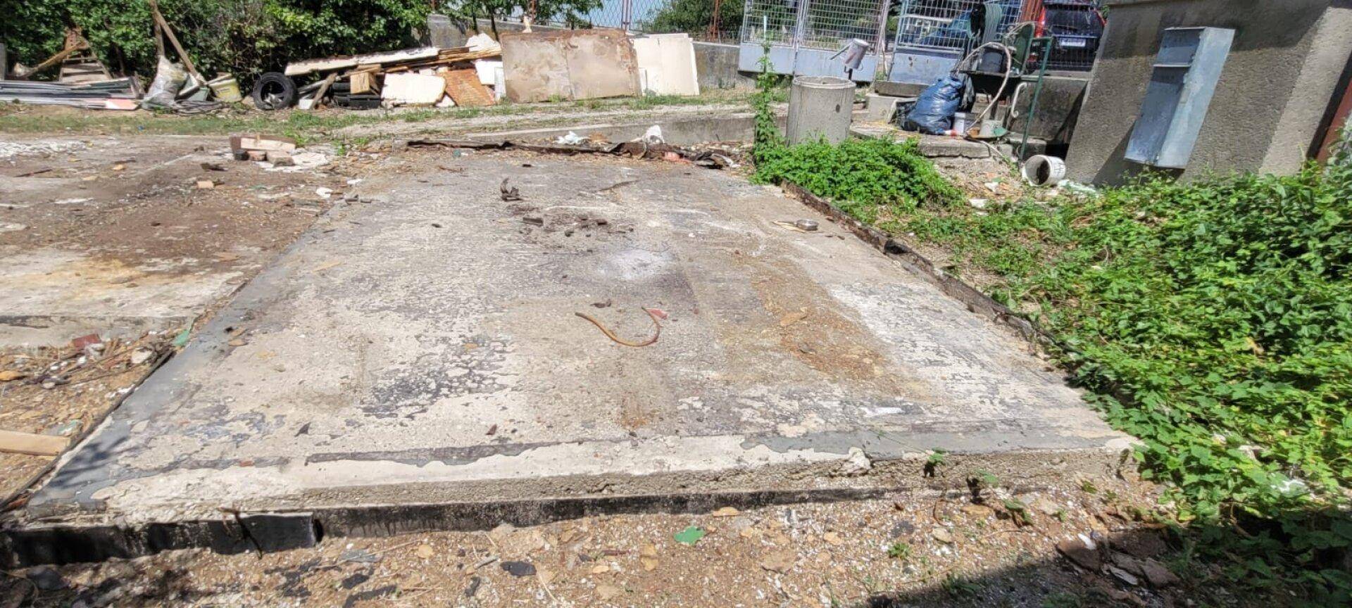 Alte Betonplatte auf einem Grundstück, umgeben von Erde und spärlicher Vegetation.