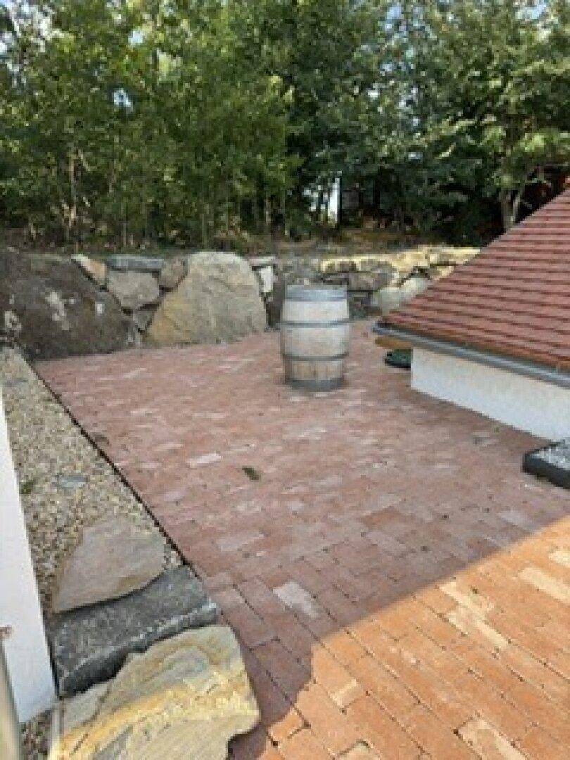 Gepflasterte Terrasse mit Natursteinmauer und einem Holzfass, umgeben von Bäumen.