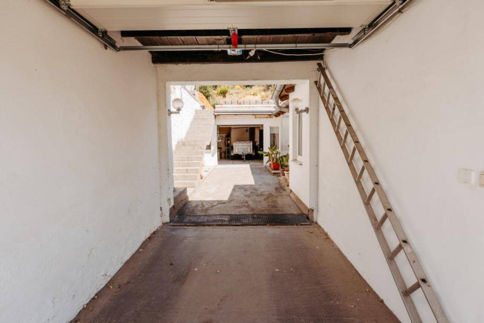 Überdachter Durchgang oder Garage mit Betonboden, der zu einem weiteren Bereich mit Treppe führt.