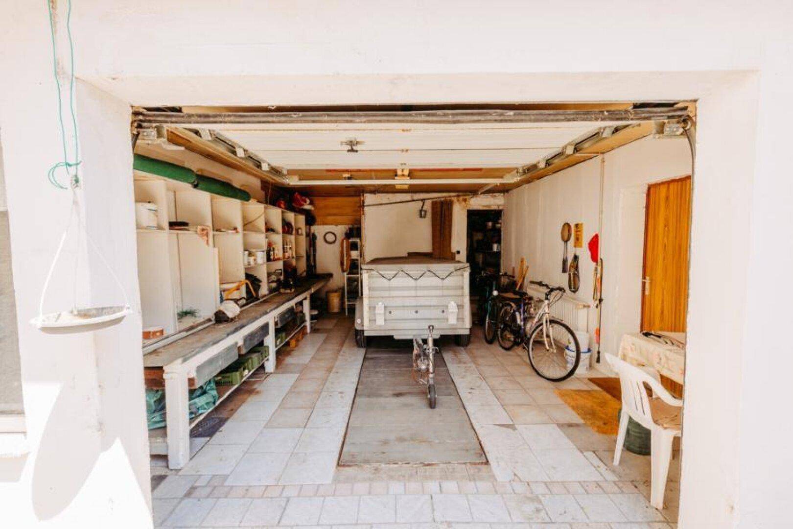 Geräumige Garage mit Fliesenboden, Abstellregalen, einem Anhänger und Fahrrädern.