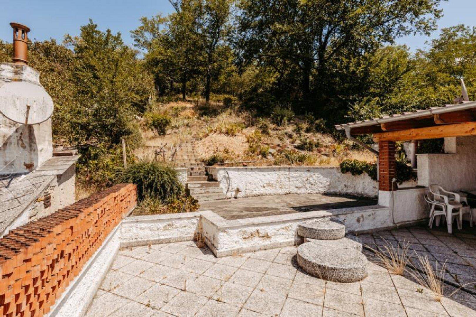 Große Außenterrasse mit Steinplatten, niedrigen Mauern und Blick auf bewaldete Hügel.
